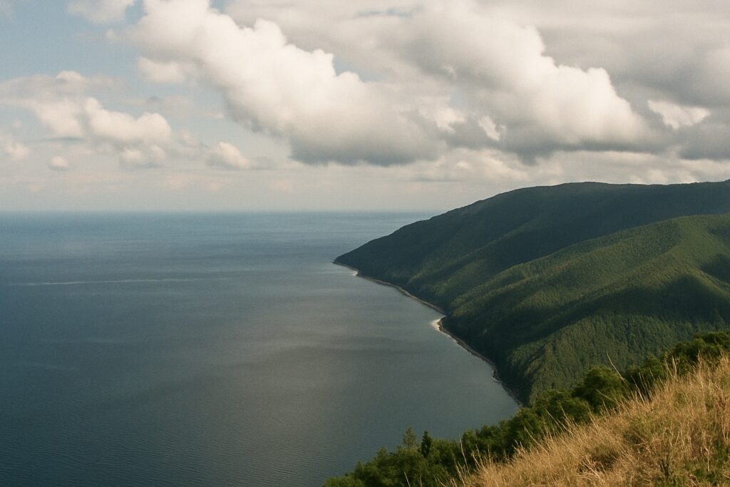 Blick von einem Hang über den weiten See und eine bewaldete Bergkette mit Wolkenschatten